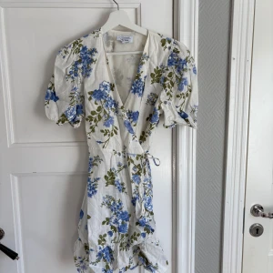 Vit blommig omlottklänning från & other stories - Söt vit omlottklänning från & other stories med blått blommönster och gröna blad. Klänningen har volangdetaljer, korta puffärmar och knyt i midjan. Perfekt för sommarens alla tillfällen!
