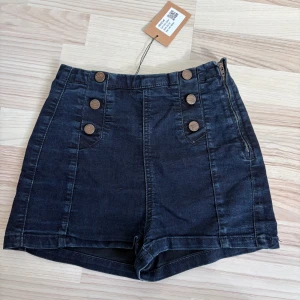 Svarta högmidjade jeansshorts - Snygga svarta högmidjade shorts från ginatricot i jeansmaterial. Shortsen har dekorativa knappar framtill och dragkedja i sidan för en cool look. Perfekta till sommaren och lätta att matcha med olika toppar.