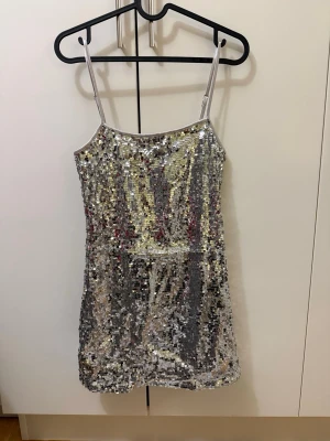 Glittrig silver paljettklänning - Säljer en kort, glittrig klänning i silver med tunna axelband och täckt av paljetter. Perfekt för dig som vill sticka ut och glänsa på festen. Klänningen har en figurnära passform och är ärmlös.