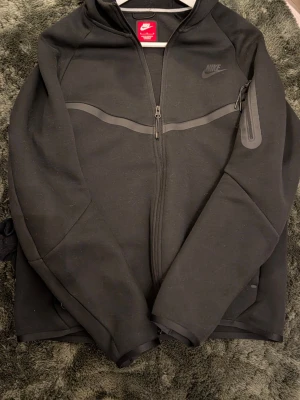 Svart Nike Tech - Snygg svart hoodie från Nike med hel dragkedja framtill och diskret Nike-logga på bröstet. Hoodien har en huva, en ficka med dragkedja på ärmen och är gjord i ett mjukt, stretchigt material. Perfekt för en sportig och avslappnad stil.