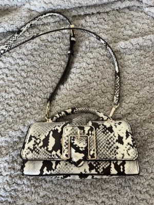 Vit och svart axelremsväska i ormmönster från Zara - Snygg handväska i vit och svart ormmönstrad faux läder med guldfärgade detaljer och justerbar axelrem. Väskan har ett stort spänne framtill och magnetlås. Perfekt för dig som gillar statement-accessoarer och trendiga detaljer.