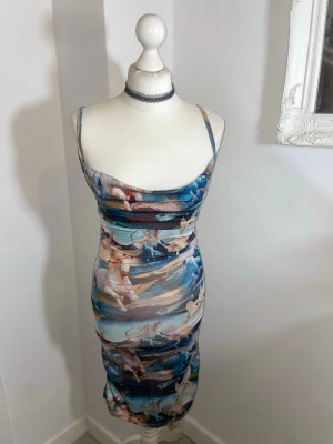 Mönstrad långklänning med hästar - Unik långklänning med smala axelband och konstnärligt hästmönster i blå, beige och bruna toner. Klänningen har figurnära passform och är tillverkad i ett stretchigt syntetmaterial som sitter snyggt på kroppen. Perfekt för dig som vill sticka ut med en cool och kreativ look.