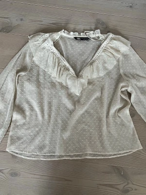 Vit blus med volang från Zara - Söt vit blus från Zara med transparent tyg och diskret prickstruktur. Blusen har en djup V-ringning med volangdetaljer och knytband vid halsen. Långa ärmar i lätt och luftig kvalitet, perfekt för en romantisk look.