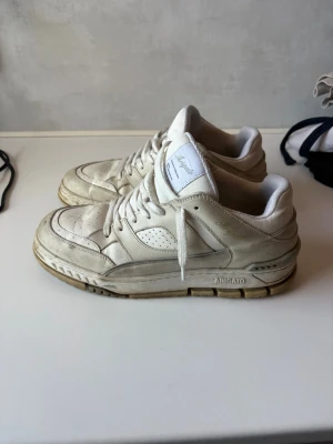 Vita sneakers från Axel Arigato - Stilrena vita sneakers från Axel Arigato med lågt skaft och klassisk siluett. Skorna har perforerade detaljer på sidan och tåpartiet, samt en robust sula i gummi. Tillverkade i läder med vita skosnören och diskret logga på sidan. Passar perfekt med alla byxor, speciellt mjukis, jeans eller shorts med Nike strumpor.