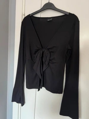 Svart långärmad topp med knyt - Trendig svart långärmad topp med djup v-ringning och snygg knytning framtill. Modellen är croppad och har en smickrande passform. Perfekt att styla med höga jeans eller kjol för en cool look.