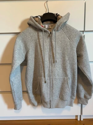 Grå zip hoodie från Burberry - Snygg grå zip hoodie från Burberry med klassiskt rutigt foder i huvan. Hoodien har dragsko i huvan, två fickor framtill och ribbade muddar vid ärmar och nederkant. Tillverkad i mjukt bomullsmaterial som är perfekt för en avslappnad stil.
