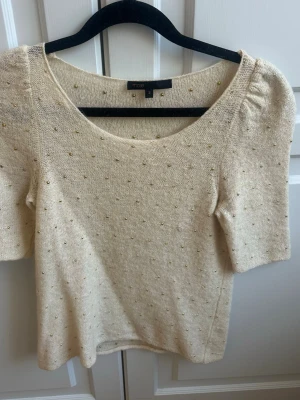 Beige topp med gulddetaljer från Maje - Supersöt beige topp från Maje med små guldfärgade pärlor över hela plagget. Toppen har korta ärmar, rund halsringning och är stickad i ett mjukt material. Perfekt för dig som vill ha något unikt och stilrent i garderoben.