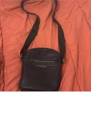 Svart axelväska från Valentino - Snygg svart axelväska äkta från Valentino med justerbar axelrem och silverfärgade detaljer. Väskan har en dragkedjeficka framtill och är tillverkad i ett tåligt konstläder med struktur. Perfekt för att bära det viktigaste på ett stilrent sätt. Priset kan diskuteras och mer ditaljer och bilder på väskan