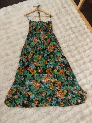 Blommig långklänning med smock - Snygg långklänning med färgglatt blommönster sopar golvet! Klänningen har smock upptill, tunna axelband och en flowy passform. Perfekt för sommarens varma dagar och har gröna, gula, orangea och blå toner i mönstret.