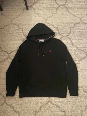 Svart hoodie från U.S. Polo Assn - Svart hoodie från U.S. Polo Assn med röd broderad logga på bröstet. Klassisk huva med dragsnören och guldfärgade detaljer. Tillverkad i mjukt bomullsmaterial och har en känguruficka framtill. Perfekt för en avslappnad och stilren look.