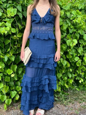 Marinblå balklänning med volanger - Säljer en marinblå långklänning med v-ringning, spetsdetaljer i midjan och flera lager volanger. Klänningen har smala axelband och en feminin, flowy siluett. Perfekt till balen! Den är endast använd en gång och har inga defekter, köptes för ca 1500kr. Jag är 165 cm för referens