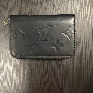 Plånbok  - Ny, liten svart plånbok med silvrig dragkedja och mönster på. Fack för kort mm.. Bara legat i plastpåsen den kom i och box, sen sen köptes. Kommer med box också, om man vill ha. Ca 10.5 cm och 7 cm hög.