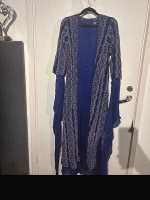 Blå klänning xs/s - Snygg blå morgonrock i lång modell med vita och guldiga broderade detaljer längs kanterna. Morgonrocken har långa ärmar och är gjord i ett tunt, flytande tyg som ger en elegant känsla. Perfekt för att chilla hemma i stil.