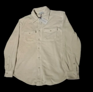 Beige overshirt från Sandö - Aldrig använd med tagg kvar. Org. Pris 700:- . Stilren beige overshirt från Sand med klassisk krage, två bröstfickor med lock och knappar framtill. Tillverkad i mjukt material som ger en avslappnad men ändå snygg look. Perfekt att bära över en t-shirt eller hoodie.