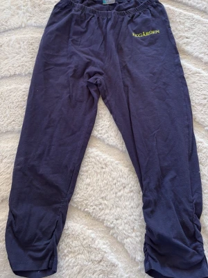 Mörkblå mjukisbyxor från Trofé - Mörkblå mjukisbyxor från Trofé med resår i midjan och tryckt text 'TRÄDGÅRDEN' i gult på ena benet. Tillverkade i mjukt bomullsmaterial och har rynkade detaljer vid bensluten. Perfekta för chill dagar hemma eller när du vill vara bekväm.