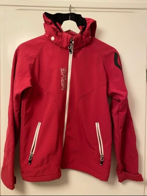 Röd vindjacka från Wave Protect - Snygg röd vindjacka från Wave Protect med vit dragkedja och två vita fickor framtill. Jackan har huva, svart innerfoder i fleece och en cool detalj på ärmen. Perfekt för blåsiga dagar och har en sportig look.