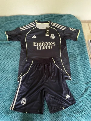 Real Madrid svart matchställ Adidas - Snyggt svart matchställ från Real Madrid och Adidas. Setet består av en kortärmad tröja och shorts med vita detaljer och klubbmärke på både tröja och byxa. Materialet är lätt och andas, perfekt för fotboll eller träning. Sponsoremblem och Adidas-logga framtill.