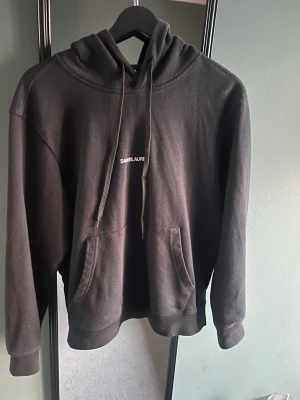 Saintlaurent Hoodie - Storlek S men passar xs.Svart hoodie från Saintlaure med klassisk känguruficka och justerbar huva med snören. Minimalistisk design med liten vit logga på bröstet. Tillverkad i mjukt material som känns skönt mot huden. Perfekt för en avslappnad och stilren look. 