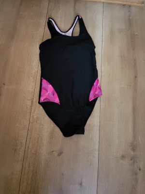 Svart baddräkt med rosa detaljer - Snygg svart baddräkt med sportig rygg och rosa mönstrade partier på sidorna och upptill på ryggen. Klassisk modell med rundad halsringning och breda axelband. Perfekt för simträning eller strandhäng.