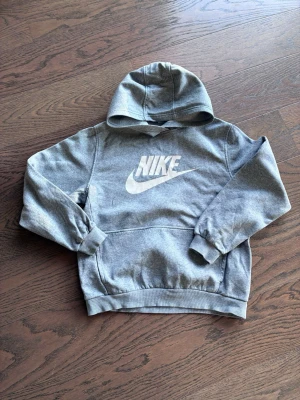 Grå hoodie från Nike med logga - Snygg grå hoodie från Nike med stor vit logga på bröstet. Klassisk känguruficka framtill och ribbade muddar vid ärmar och nederkant. Tillverkad i mjukt sweatshirtmaterial som är skönt att ha på sig. Perfekt för en avslappnad streetwear-look.