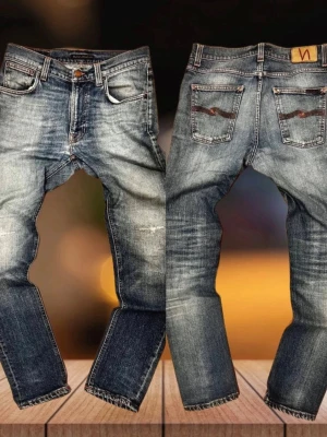 Blå slitna jeans från Nudie Jeans - Säljer ett par blå jeans från Nudie Jeans med snyggt slitna detaljer och klassisk femficksmodell. Jeansen har raka ben, kontrastsömmar och Nudie-logga på bakfickan. Tillverkade i kraftig denim med tvättad finish för en cool look.