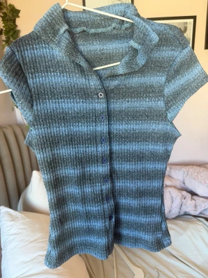 Gråblå ribbad topp med knappar - Kortärmad ribbad topp i gråblått med horisontella ränder och knappar framtill. Har en liten krage och figurnära passform. Perfekt till jeans eller kjol för en chill look. märket står inte men tippar på storlek s/xs