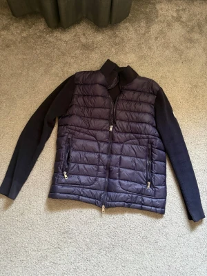 Mörkblå dunväst från Moncler - Snygg mörkblå dunväst från Moncler med stickade ärmar och två dragkedjeförsedda fickor framtill. Västen har hög krage och klassisk Moncler-logga på ärmen. Perfekt för lager-på-lager och streetstyle. Ingen aning om den är äkta då den är köpt från plick men det är ju också därav priset, vi kan även diskutera priset!