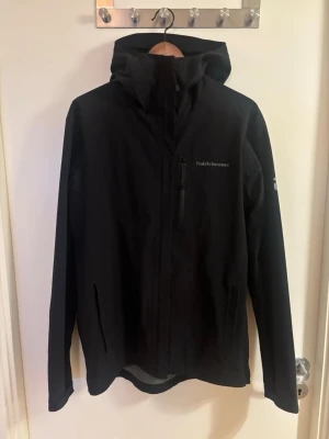 Peak Performance Jacka - Snygg svart vindjacka från Peak Performance med huva och dragkedja framtill. Jackan har en bröstficka med dragkedja och diskret logga på bröstet samt ärmen. Tillverkad i ett lätt och slitstarkt material som står emot vind.