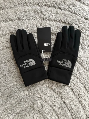 Svarta handskar från The North Face - Snygga svarta handskar från The North Face med logga i vitt på ovansidan. Handskarna har en stilren design och är tillverkade i ett  syntetmaterial som passar perfekt för kyliga dagar.