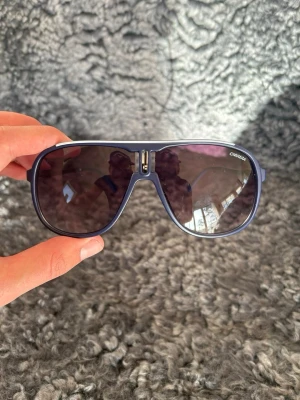 Mörkblå pilotsolglasögon Carrera - Snygga mörkblå pilotsolglasögon från Carrera med bred båge och mörka tonade glas. Klassisk aviatorform med metalliga detaljer vid näsryggen. Perfekta för dig som vill ha en cool och tidlös stil i solen.