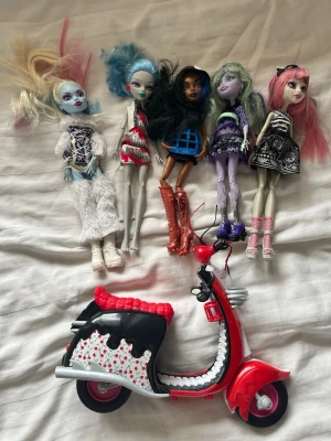 Monster High dockor och röd scooter - Fem Monster High dockor med olika färgglada kläder och hår i blått, svart, grönt och rosa. Dockorna har detaljerade outfits och höga stövlar. Medföljer gör en röd och svart leksaksscooter med vita och röda detaljer samt hjärtformade mönster på sätet.