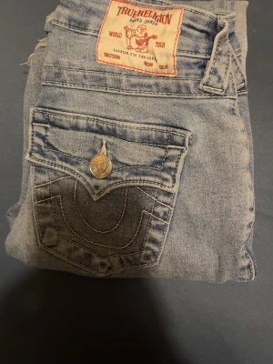 True Religion ljusblå jeans - Säljer ett par mid waist ljusblå jeans från True Religion med deras klassiska kontrastsömmar och snygga ficklock bak med stora silverfärgade knappar. Jeansen har en avslappnad passform och är tillverkade i mjuk denim. Perfekta för en cool och avslappnad stil. Ordinarie pris 1500kr säljer för 1000kr