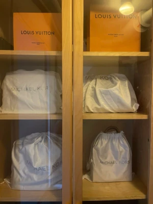 Svart handväska från Michael Kors - Snygg svart handväska från Michael Kors med guldigt MK-hänge och dubbla handtag. Väskan har en klassisk rektangulär form och är tillverkad i slätt konstläder. Perfekt för dig som gillar stilrena accessoarer med exklusiv känsla.