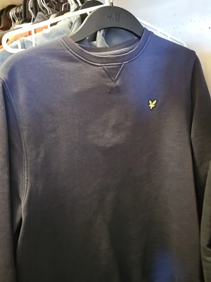 Mörkblå sweatshirt från Lyle & Scott - Mörkblå sweatshirt från Lyle & Scott med klassisk rund halsringning och det ikoniska gula örnlogot på bröstet. Tröjan har långa ärmar och ribbade muddar vid ärmslut och nederkant. Perfekt för en avslappnad och stilren look.