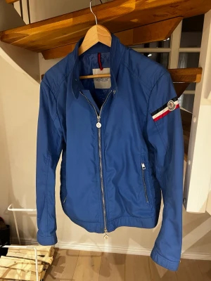 Moncler Courchlevel - Blå vindjacka från Moncler, modellen Courchlevel. Jackan har två fickor med dragkedja och Moncler-logga på ärmen tillsammans med en röd, vit och blå rand. Perfekt för dig som vill ha en stilren och sportig look. Skickar självklart samma dag som köp 👍🙂