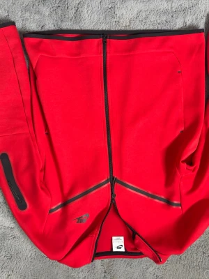 Röd Nike hoodie med dragkedja - Snygg röd hoodie från Nike med hel dragkedja framtill och svarta detaljer. Hoodien har en huva och praktiska fickor på sidorna, samt Nike-logga på framsidan. Tillverkad i mjukt material som känns skönt mot huden. Perfekt för en sportig och avslappnad stil.
