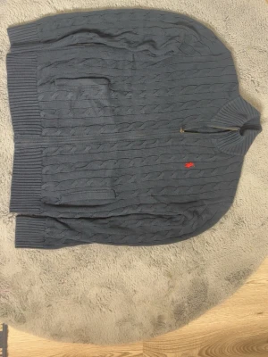 Mörkgrå kabelstickad tröja Polo Ralph Lauren - Kabelstickad mörkgrå tröja från Polo Ralph Lauren med dragkedja, hög krage och två fickor framtill. Tröjan har det klassiska röda Polo-logot på bröstet och ribbade muddar vid ärmslut och nederkant. Perfekt för lager på lager och snygg till jeans.