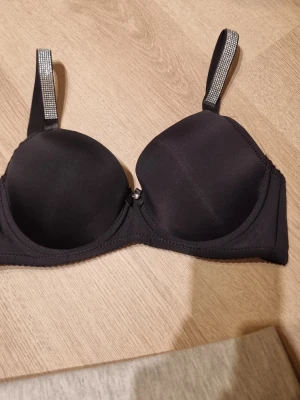Svart bh med strassband - Snygg svart bh med vadderade kupor och bygel. Axelbanden är dekorerade med glittrande strassdetaljer för extra stil. Bh:n har hakknäppning bak och släta kupor utan spets. Perfekt för dig som vill ha något enkelt men ändå glam. Storlek 75b men är liten i storleken. 
