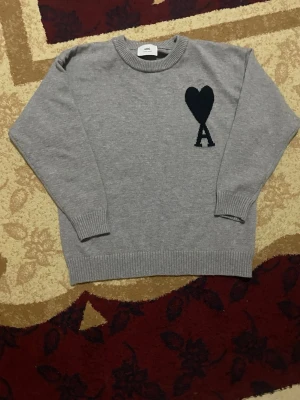 Grå stickad tröja från Ami Paris - Snygg grå stickad tröja från Ami Paris med svart hjärta och bokstaven A broderat på bröstet. Tröjan har rund halsringning, ribbade muddar och är tillverkad i mjukt material som passar perfekt till lager på lager.