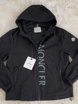 Svart vindjacka från Moncler - Snygg svart vindjacka från Moncler med huva och dragkedja framtill. Jackan har Moncler-logga på ärmen och stort tryck med Moncler på framsidan. Två fickor med dragkedja och elastiska muddar vid ärmslut. Perfekt för en trendig streetwear-look.