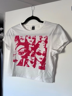 Vit baby tee med rött tryck - Säljer en vit baby tee med ett rött popkonst-tryck. Perfekt för dig som gillar grafiska motiv och streetstyle. Liten i storleken!! Passar till S/M.