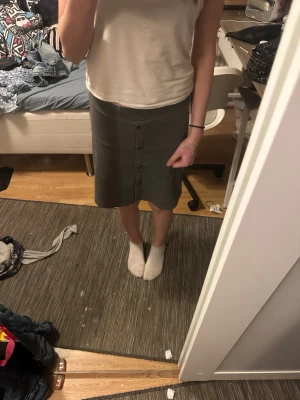 Grå kjol med knappar från Name It - Säljer en grå kjol från Name It i storlek 164. Kjolen har en rak passform, knappar framtill och är knälång. Perfekt för dig som gillar enkel och stilren design.