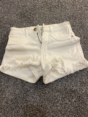 Vita fransiga shorts från Levi's - Säljer ett par vita lågmidjade shorts från gina med fransiga benslut och klassisk femficksdesign. Shortsen har knapp och dragkedja framtill samt en liten läderpatch bak i midjan. Perfekta för sommaren.