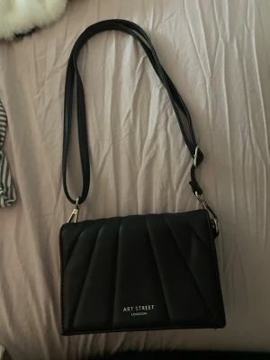 Svart axelväska från Art Street - Snygg svart axelväska från Art Street London med quiltad design och justerbar axelrem. Väskan har silverfärgade detaljer och en stilren, rektangulär form. Perfekt för dig som gillar minimalistisk och trendig stil.