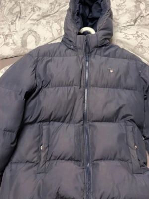 Mörkblå pufferjacka från Gant - Snygg mörkblå pufferjacka från Gant med hög krage och huva. Jackan har två framfickor med tryckknappar och dragkedja framtill. Klassisk quiltad design och liten Gant-logga på bröstet. Perfekt för kalla vinterdagar.