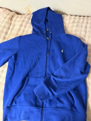 Blå hoodie från Polo Ralph Lauren - Snygg blå hoodie från Polo Ralph Lauren med dragkedja framtill och klassisk logga broderad på bröstet. Hoodien har justerbar huva med snören och två fickor framtill. Perfekt för en avslappnad och stilren look. Den har tyvärr en liten fläck på framsidan men äkta! :)