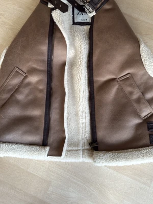 Brun fårskinnsinspirerad dunväst Zara - Snygg brun dunväst från Zara med fårskinnsinspirerat foder i vitt. Västen har två fickor framtill, dragkedja och coola spänndetaljer vid kragen och sidorna. Perfekt för lager på lager och ger en trendig look. Orginalpris cirka 700-800kr