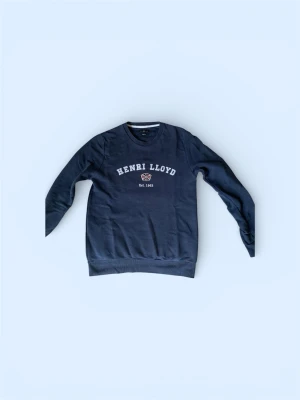 Mörkblå sweatshirt från Henri Lloyd - Mörkblå sweatshirt från Henri Lloyd med rund halsringning och tryckt logga samt texten 'Est. 1963' på bröstet. Tröjan har ribbade muddar vid ärmslut och nederkant. Perfekt för en avslappnad och stilren look.