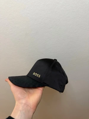 Svart keps från BOSS - Snygg svart keps från BOSS med guldfärgad logga broderad på framsidan. Skärmen har ett diskret mönster som ger extra stilpoäng. Justerbar baktill för perfekt passform. Tillverkad i ett lätt och bekvämt material.
