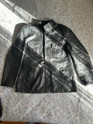 Svart skinnjacka med fickor - Snygg svart skinnjacka med fyra framfickor med lock och knapp, rak passform och ståkrage. Jackan har en stilren design med markerade sömmar och knäppning framtill. Perfekt för dig som vill ha en klassisk men cool look.